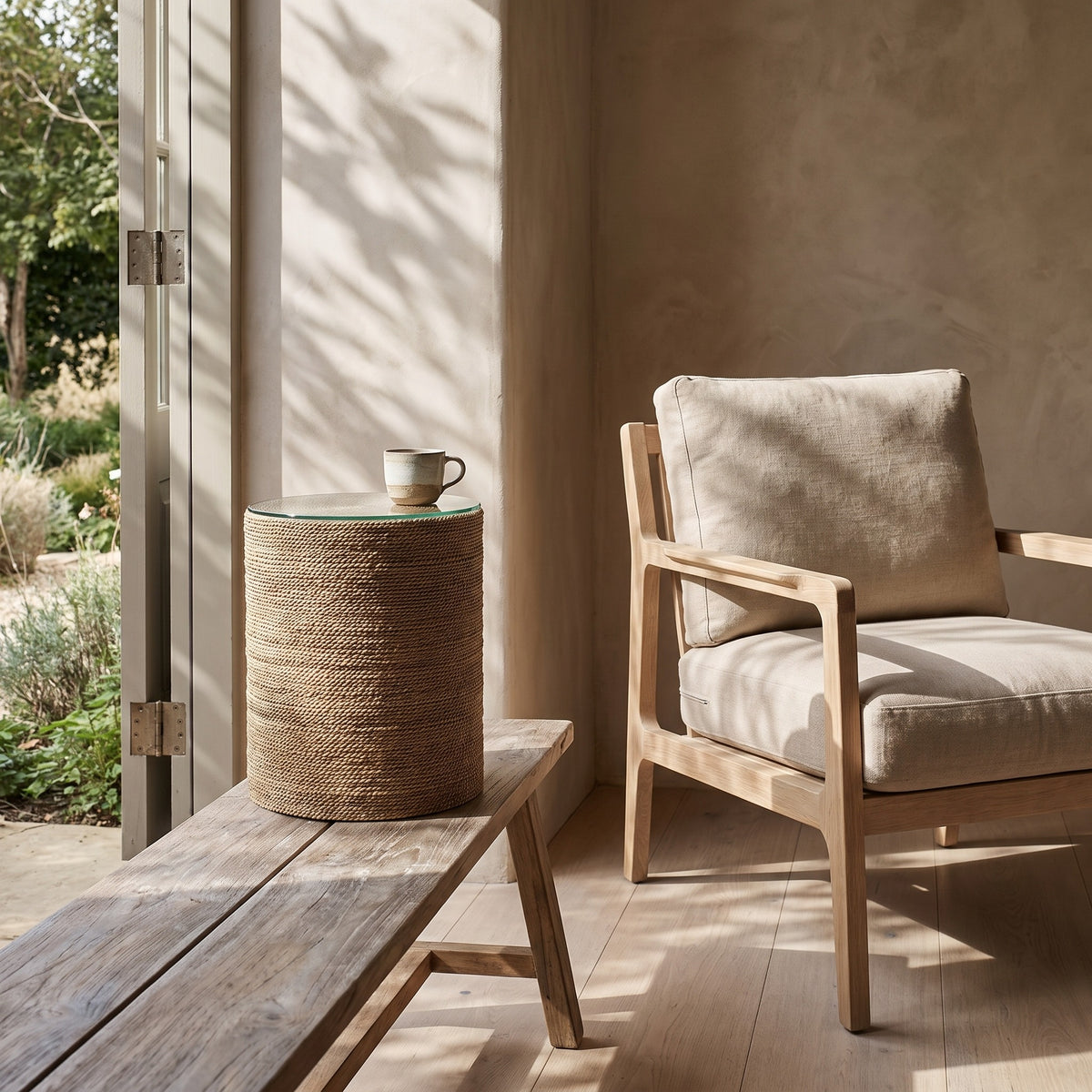 A beige cushioned chair on a light wooden floor next to a round textured side table with a glass top and a small cup on it, set outside with sunlight casting shadows from a nearby tree.