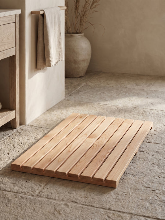 Rectangular cedar wood shower mat with slats lying on a stone floor with a towel hanging nearby.
