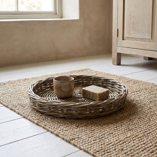 Charleston Rattan Round Tray with a cup and soap, placed on a woven mat in a room with white flooring and a cabinet.