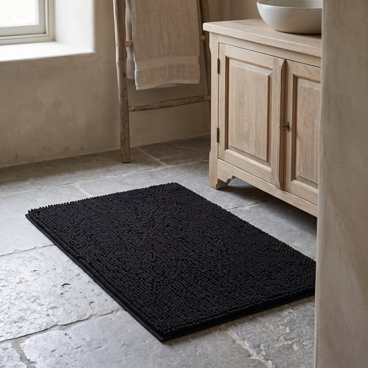 Black Chenille bath mat on stone floor next to wooden cabinet and towels.
