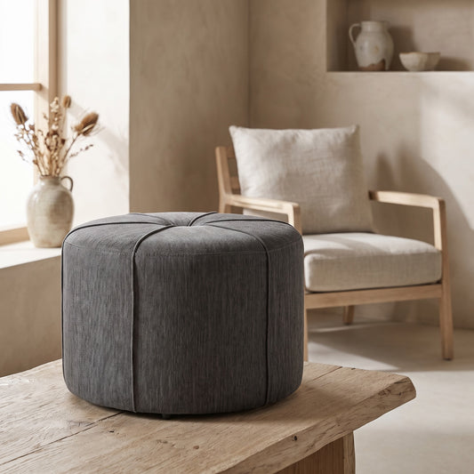 A gray ottoman with button tuft design and tonal piping on a wooden surface; natural light streaming into a room with a chair and decorative vase in the background.