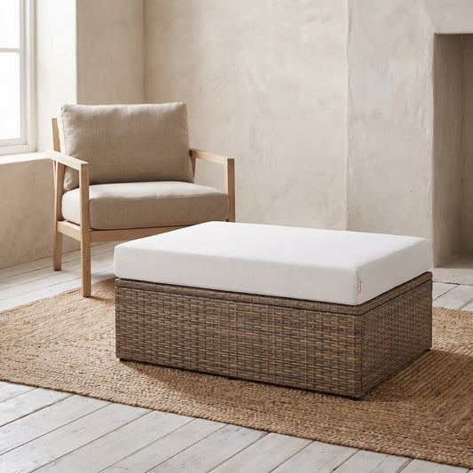 Rectangular wicker ottoman with white cushion, placed under a beige armchair on a beige rug in a minimalist room setting.