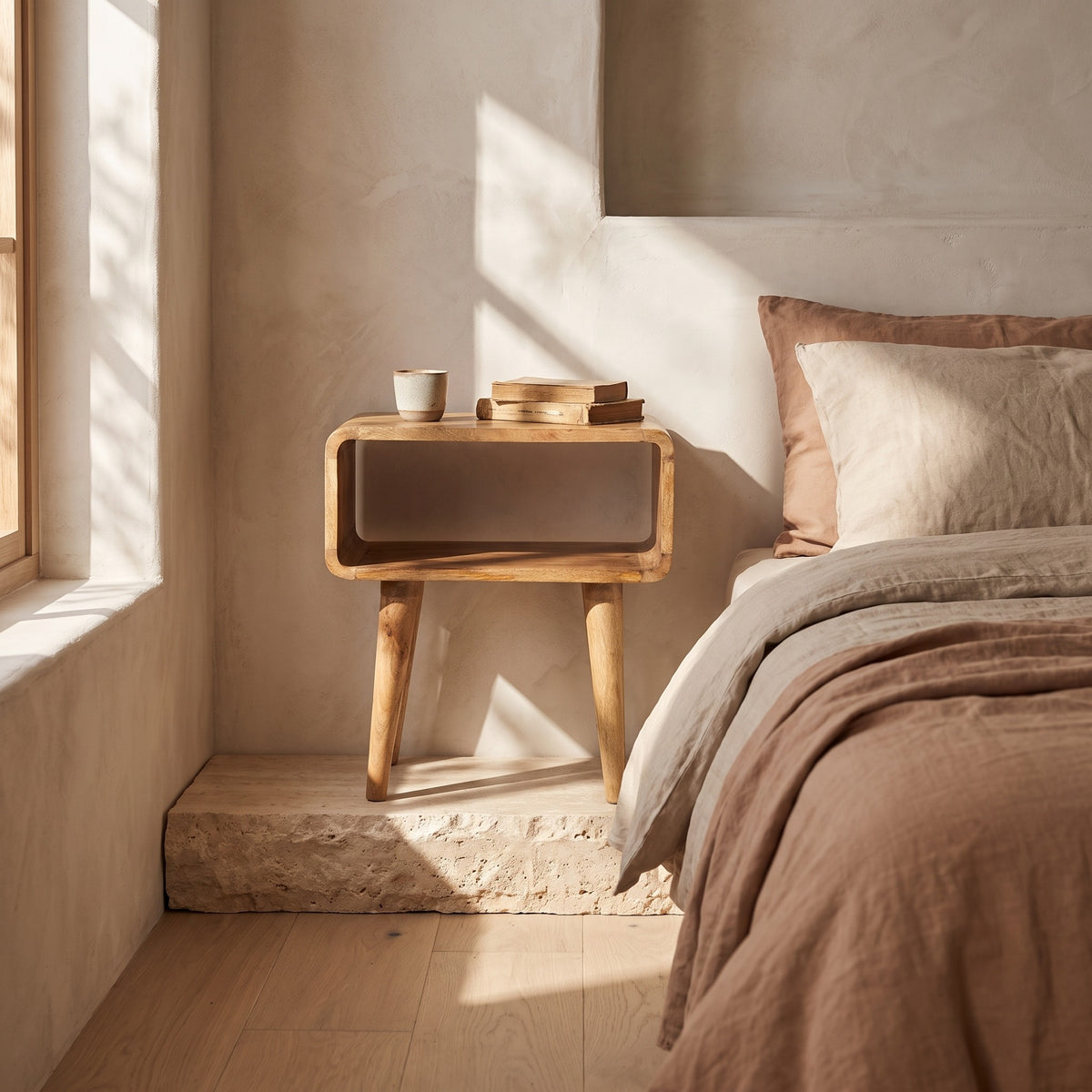 An open, curved bedside with a natural wood finish, placed next to a bed with beige bedding, under soft natural light