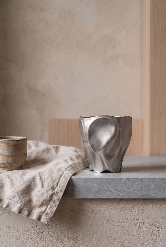 Angular, abstract silver porcelain cup on a gray stone ledge next to a beige fabric and another rounded wooden cup on the same ledge, against a textured light-colored wall with wooden paneling.