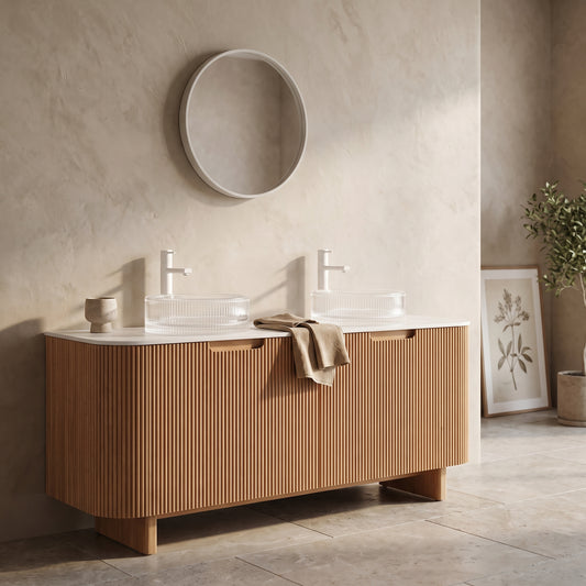Natural wood vanity with glass basins and metal faucets, placed against a textured wall with a round mirror and a potted plant nearby.