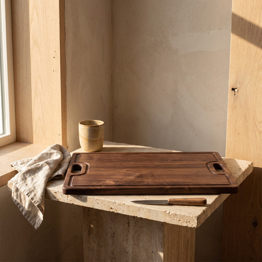 Rectangular walnut cutting board with handles, sitting on a stone table corner in natural light. Adjacent are a knife and a textile, in a minimalist setting.