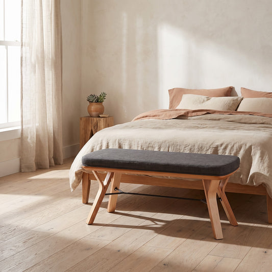 Wooden bench with gray cushion in front of a bed in a bright bedroom with wooden floors and natural light, beige curtains, and a table with plants.