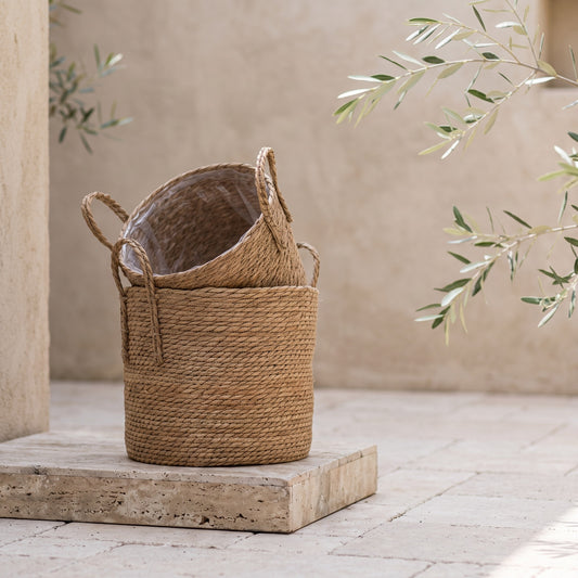 Jute basket with two handles on stone pedestal, olive branch in background, neutral wall behind, neutral sandstone floor