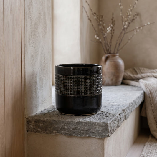 Black ceramic pot with grid pattern on a stone ledge.