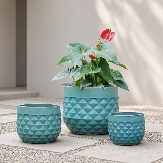Set of 3 teal ceramic planter pots with geometric pattern, one containing a plant on a paved area with a light wall in the background.