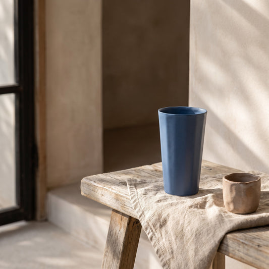A blue handmade porcelain pint cup on a wooden bench beside a window, natural setting with sunlight casting shadows.
