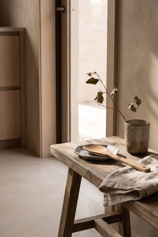 A table with a wooden spoon, a porcelain spoon rest, a ceramic vase with branches, and a folded towel, in a minimalistic setting