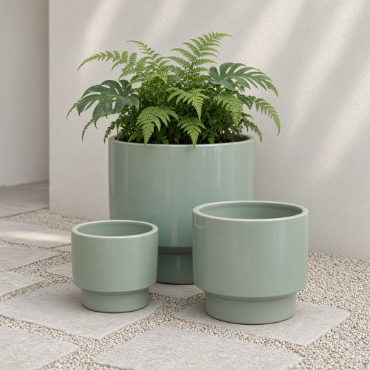 Three light green ceramic planters of varying sizes, one holding a green plant, on a tiled flooring against a plain wall.