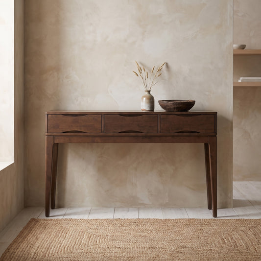 A brown wooden console table with three drawers, placed in a minimalistic room with beige walls and a natural rug on the floor.