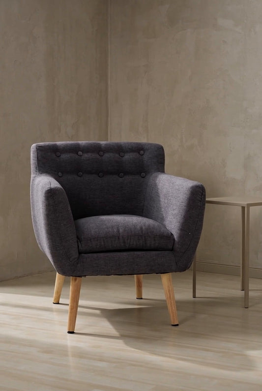A grey tufted accent armchair with wooden legs in a minimalist room with light wood flooring and a plain wall