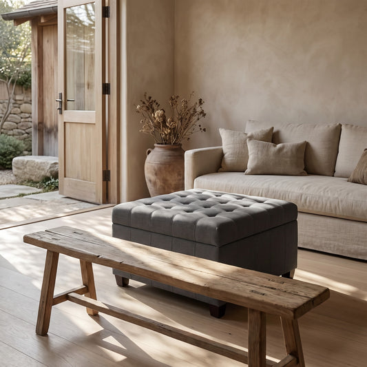 A contemporary ottoman with a tufted top in a dark gray color, placed in front of a natural beige sofa with brown textured pillows, next to a rustic wooden bench and a terracotta pot with dried flowers in a wabi-sabi home setting
