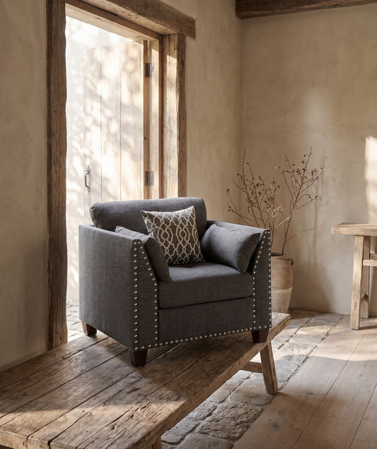 Comfortable chair in light charcoal gray with studded edges and three coordinating cushions, set in a rustic room with wooden flooring and natural light filtering through a door, next to a bare branch in a pot and a wooden bench.