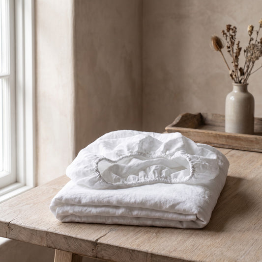 Folded white linen fitted sheet on a wooden table with a vase of dried flowers in the background.