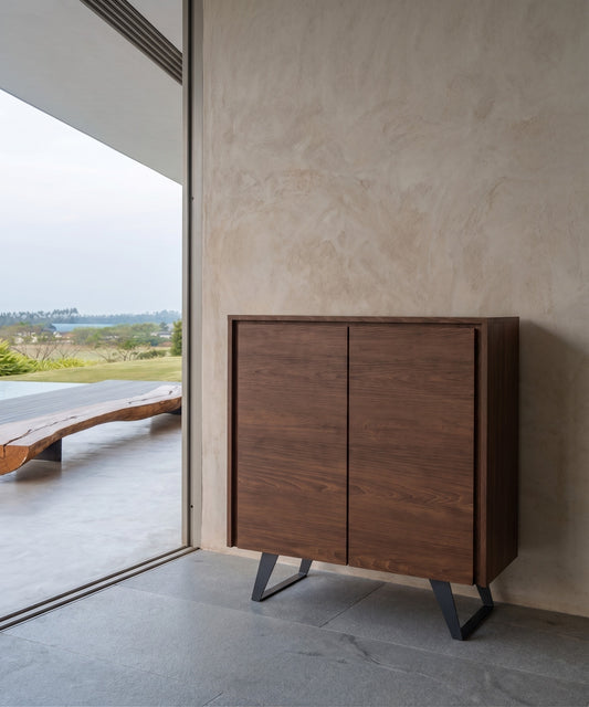 A dark wooden cabinet with sleek black metal legs, positioned in an indoor space with a view of an open outdoor area featuring a wooden bench and a scenic landscape through a sliding door.