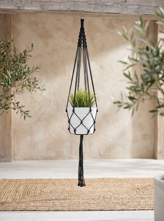 Black macrame plant hanger with white ceramic pot, hanging under wooden beam with olive leaf plant in background.