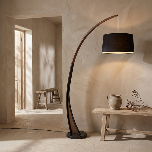 A floor lamp with an arched walnut wood frame, marble base, and linen drum shade in a minimalist room with textured walls and a wooden bench.