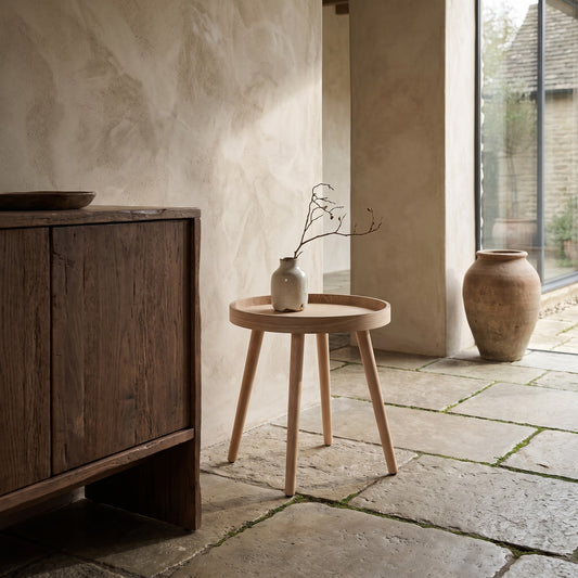 Simple wooden side table with a vase on top inside a serene, natural setting with stone flooring and rustic furniture in the background.