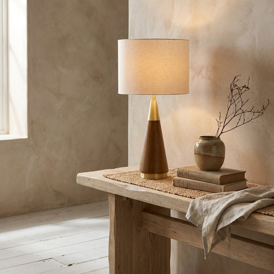 A Mid-Century Triangle Table Lamp with Espresso Wood base, Antique Brass detail, and white linen shade sits on a natural wooden table with books, a vase, and a cloth.