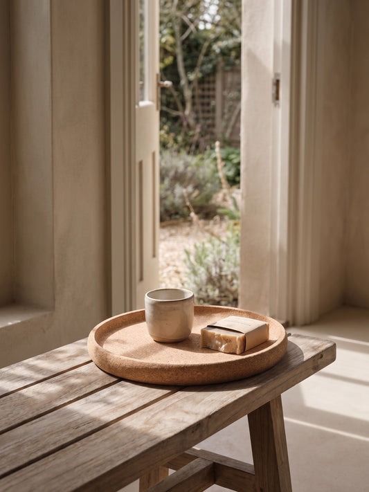 Minimal cork tray with a mug and soap on a wooden bench in front of a door leading to a garden.