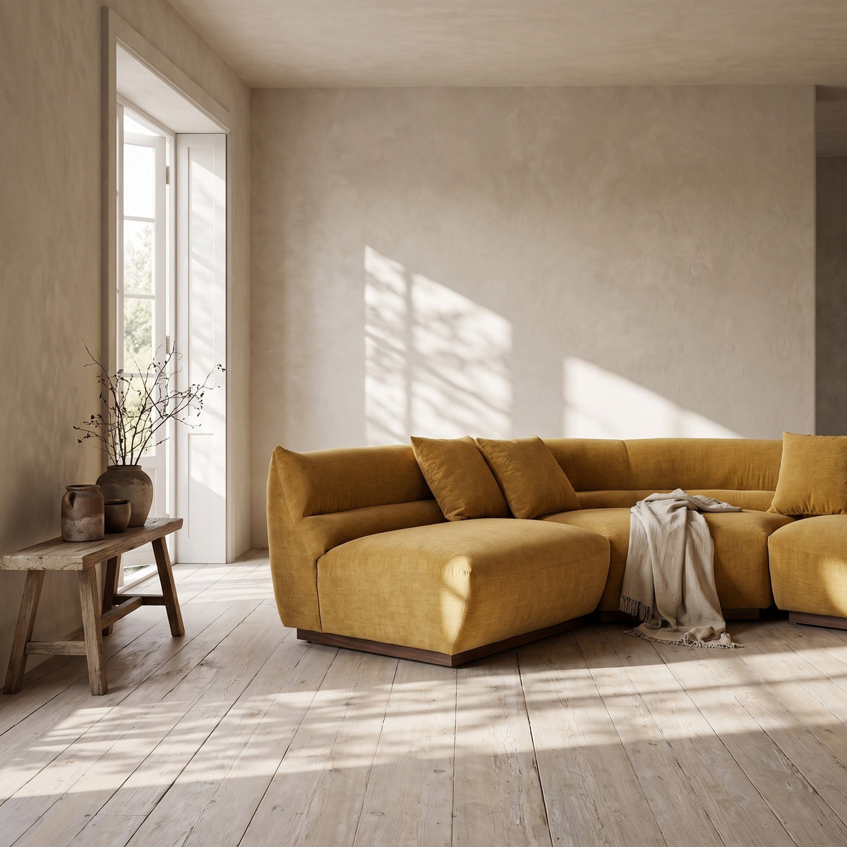 A mustard yellow sectional sofa placed in a room with natural light and wooden flooring; cushions visible; wooden side table with vase of branches beside the sofa;