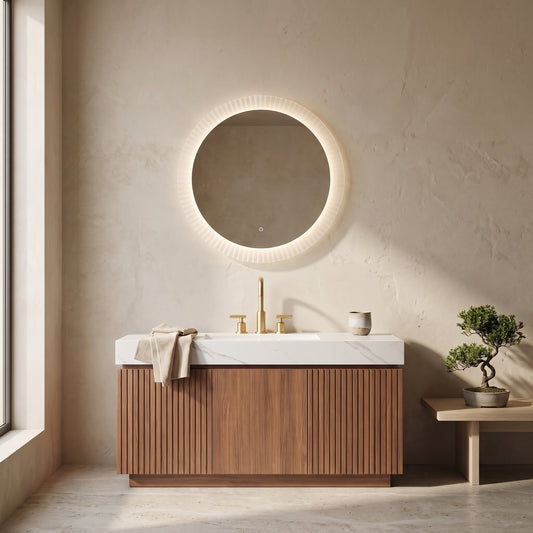 Floating vanity with wooden slats and white countertop in a minimalistic bathroom setting. Gold-colored faucets and a round illuminated mirror with a light pink/white border are visible.