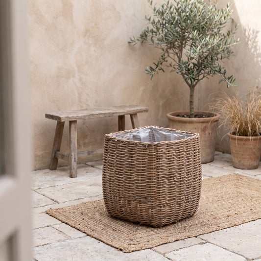 Natural wicker floor planter basket with liner on a woven mat, set against a rustic stone wall with other potted plants.