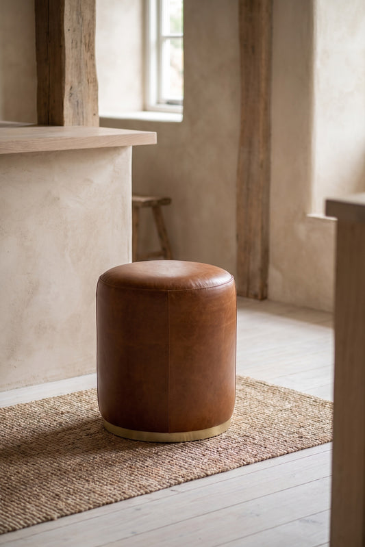 Brown round ottoman stool with leather finish on a natural fiber rug in a minimalist room with wooden beams and muted tones