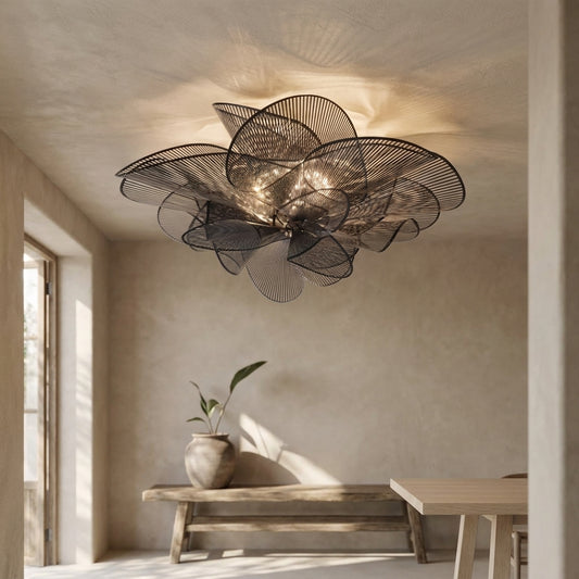 A modern black metal chandelier with petal-like details hangs from a ceiling in a minimalistic room with a wooden bench and vase.