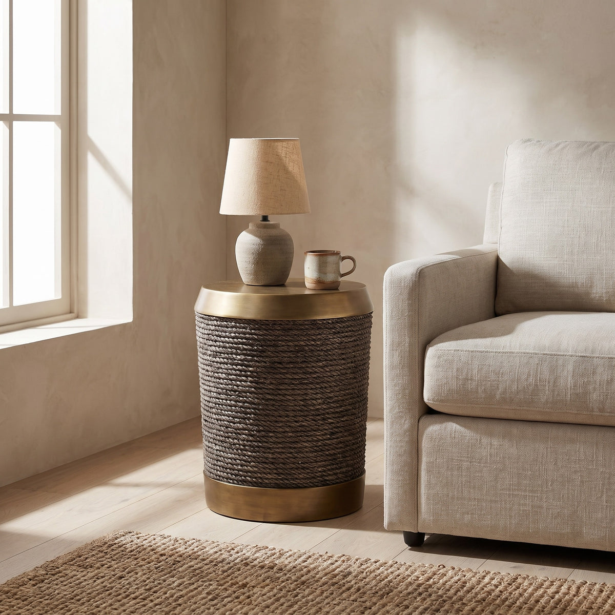 A round side table with braided rope exterior and gold base, featuring a small lamp and a mug. Beige wall background, light wood floor, beige rug, beige armchair.