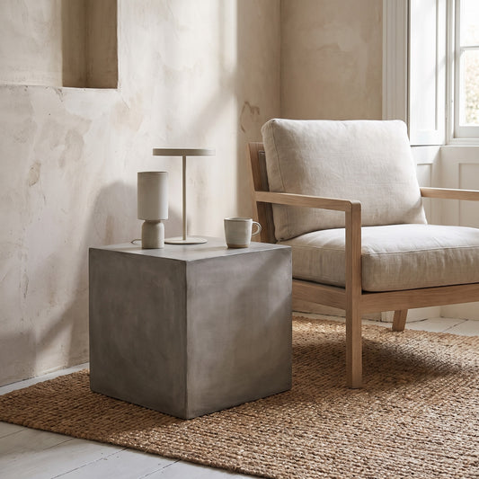 Concrete cube side table with layered lamp and mug, placed in a room with a neutral-colored chair and rug.
