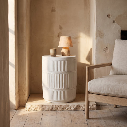 White ribbed cylindrical table with a small lamp and cup on it. Nearby, a beige armchair with a wooden frame sits on a wooden floor against a textured wall.