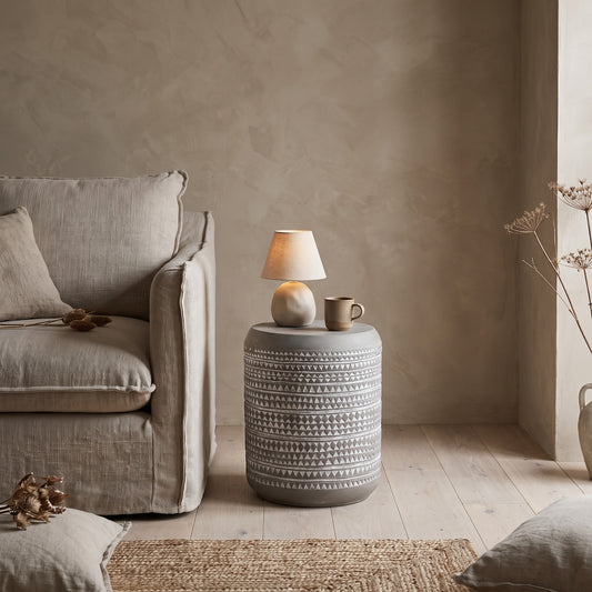 A cylindrical side table in a white and gray pattern with a lamp and mug on it, placed in a room with a beige couch and light-colored floor.