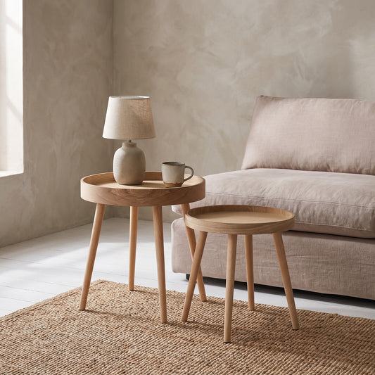 Two round wooden side tables with simple legs, one holding a lamp and a cup, placed on a textured rug in a minimalist room with a light sofa and beige wall.