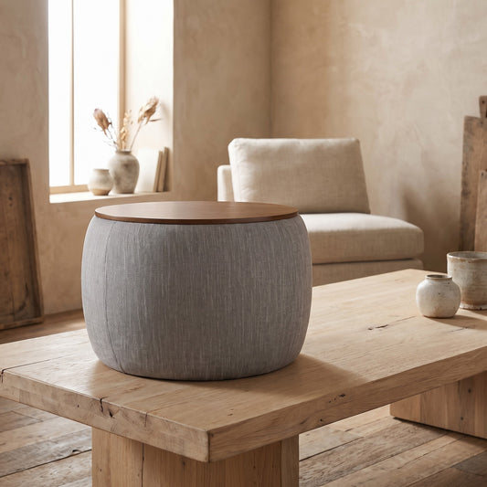 A round storage ottoman with woven heathered fabric cover and wooden top, placed on a wooden table in a minimalist living room setting.