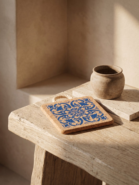 Cork trivet with blue intricate pattern on a wooden bench with a ceramic bowl in the background.