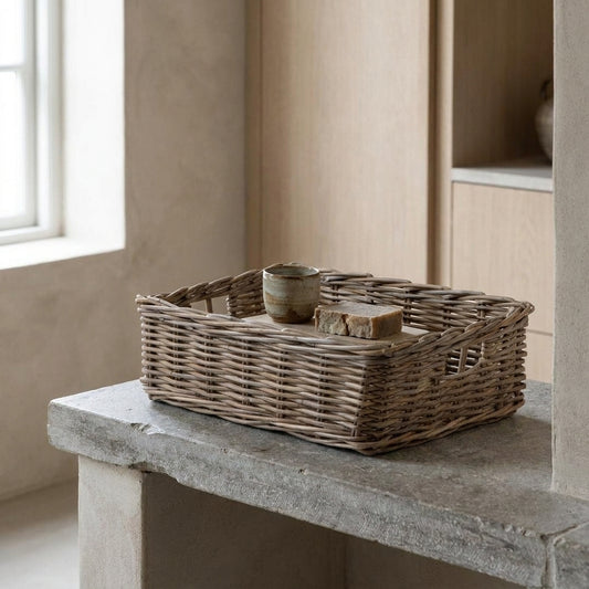 A rustic rattan rectangle basket tray holding a cup and a piece of bread on a stone bench against a minimalist wall.