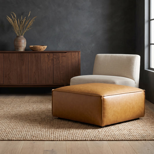 A tan vegan leather ottoman in front of a beige armchair on a textured rug with wooden floor and dark wall.