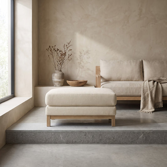A neutral-toned fabric ottoman with wooden legs, placed in a minimalist living space next to a window and a couch.
