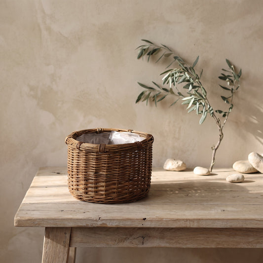 A rustic round wicker planter with a white liner sits on a wooden table in front of a light beige wall with a leafy branch and rocks in the background.