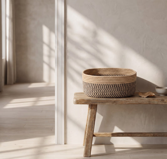 A woven jute and cotton basket with a simple pattern sits on a wooden bench, showcasing natural sunlight streaming through a window in a minimalistic room with light-colored walls.