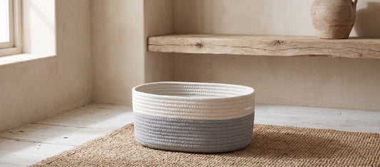White rope basket with grey ombre trim sits on a woven mat in a minimalist room with wooden shelves and a window.