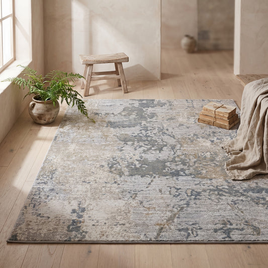 Beige rug with abstract design in shades of grey and blue on wooden floor, minimalist setting with stool and greenery.