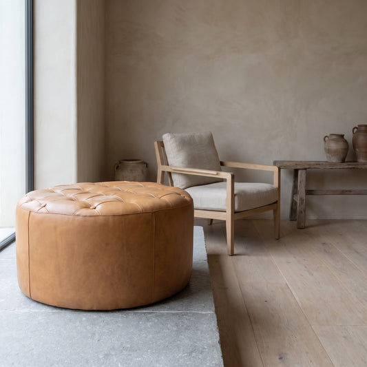 Light tan round ottoman with tufted detail, placed in a minimalist room with white walls and wooden flooring.