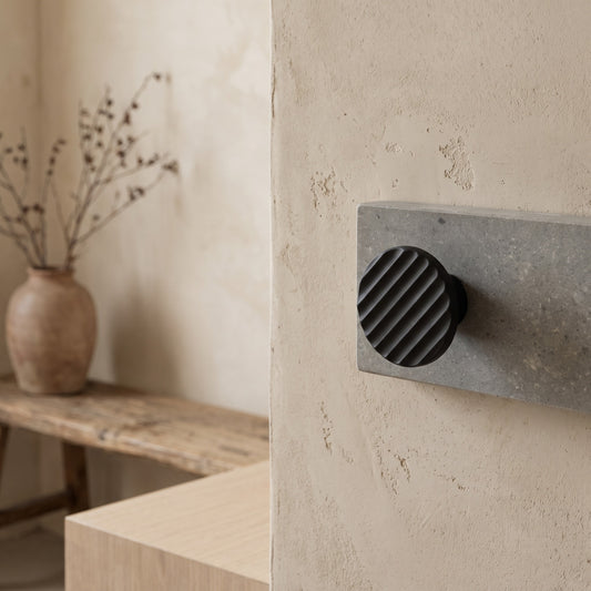 Concrete coat hook with fluted design attached to textured wall in minimalist room with vase of dried branches on a wooden shelf.