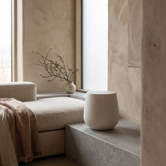 A small, round, matte off-white ceramic side table on a concrete surface near a window in a minimalist room.